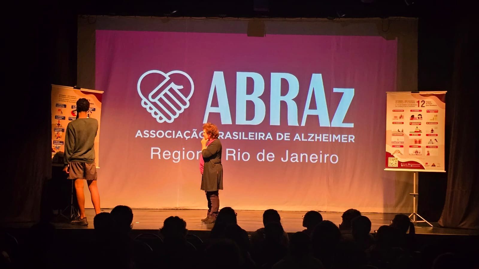 Mediadora da ABRAz falando ao microfone diante do painel da associação