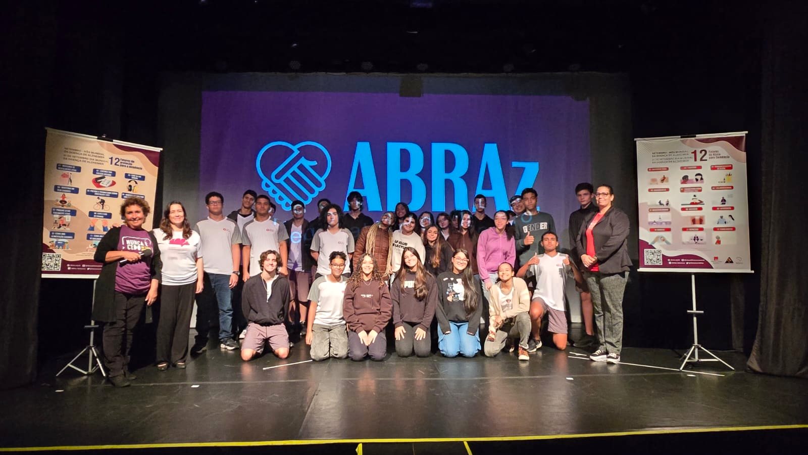 Grupo de jovens no palco do teatro em frente ao painel da ABRAz, ao lado dos banners informativos sobre a Doença de Alzheimer